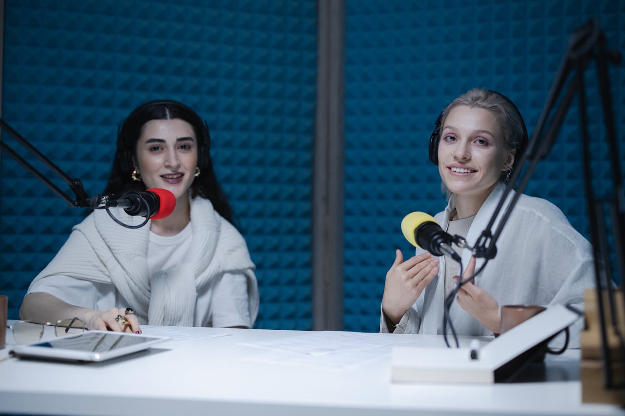 who-we-are Two women podcasters engaging in discussion inside a modern recording studio with microphones.