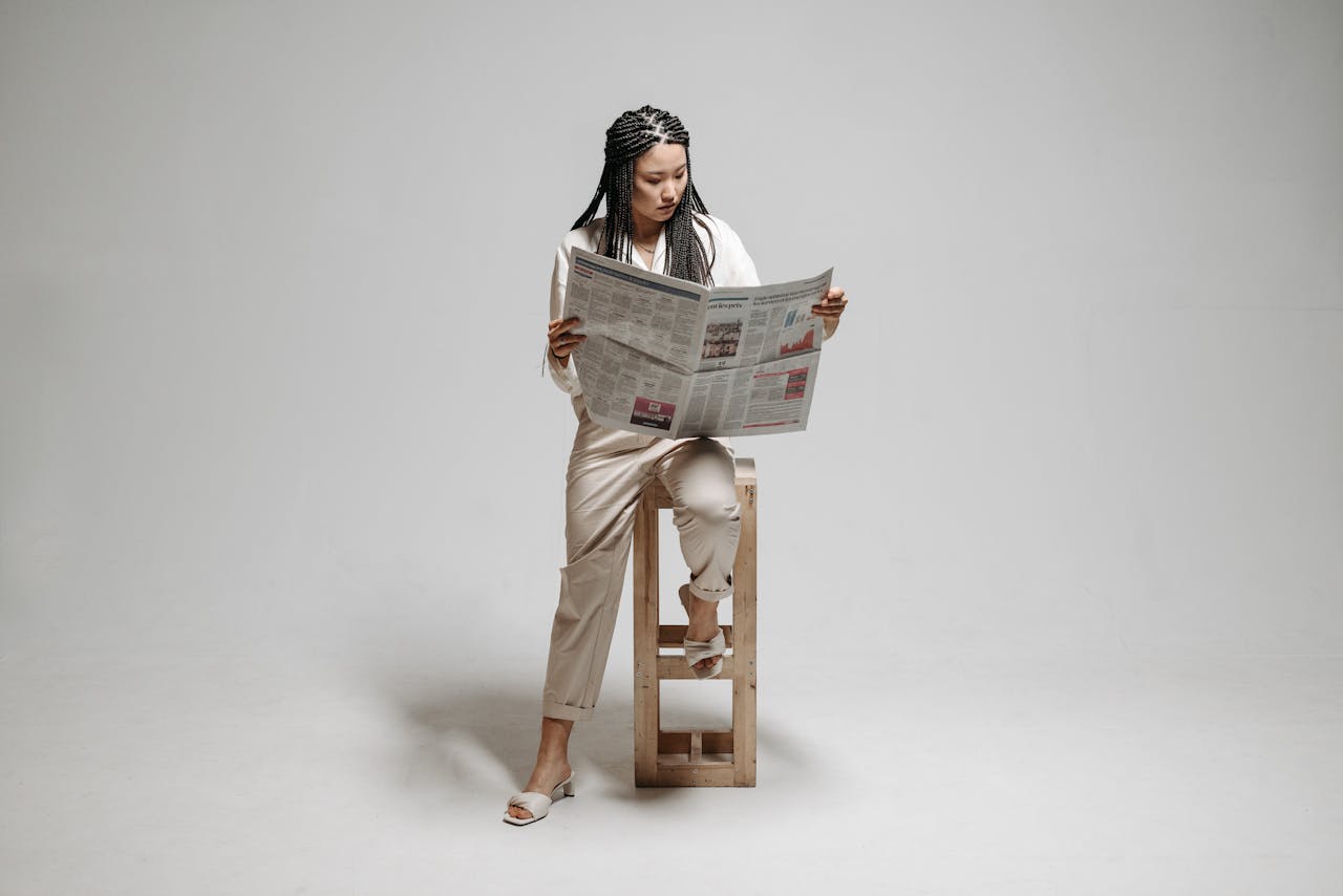 services-04 A woman sits on a stool with a braided hairstyle, reading a newspaper in a minimalistic studio.